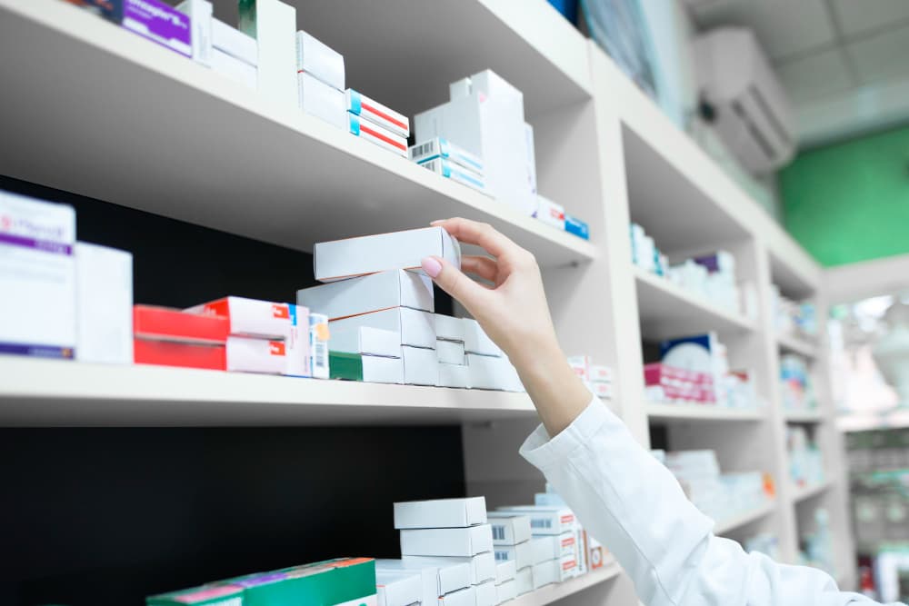 La main d'un pharmacien se tend vers une boîte de médicaments sur l'étagère d'une pharmacie garnie de divers emballages de médicaments.