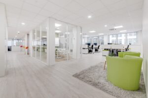 Espace de bureau moderne avec des salles de réunion en verre, des bureaux blancs, des chaises vertes et un tapis gris sur un sol en bois clair, avec de grandes fenêtres apportant de la lumière naturelle.