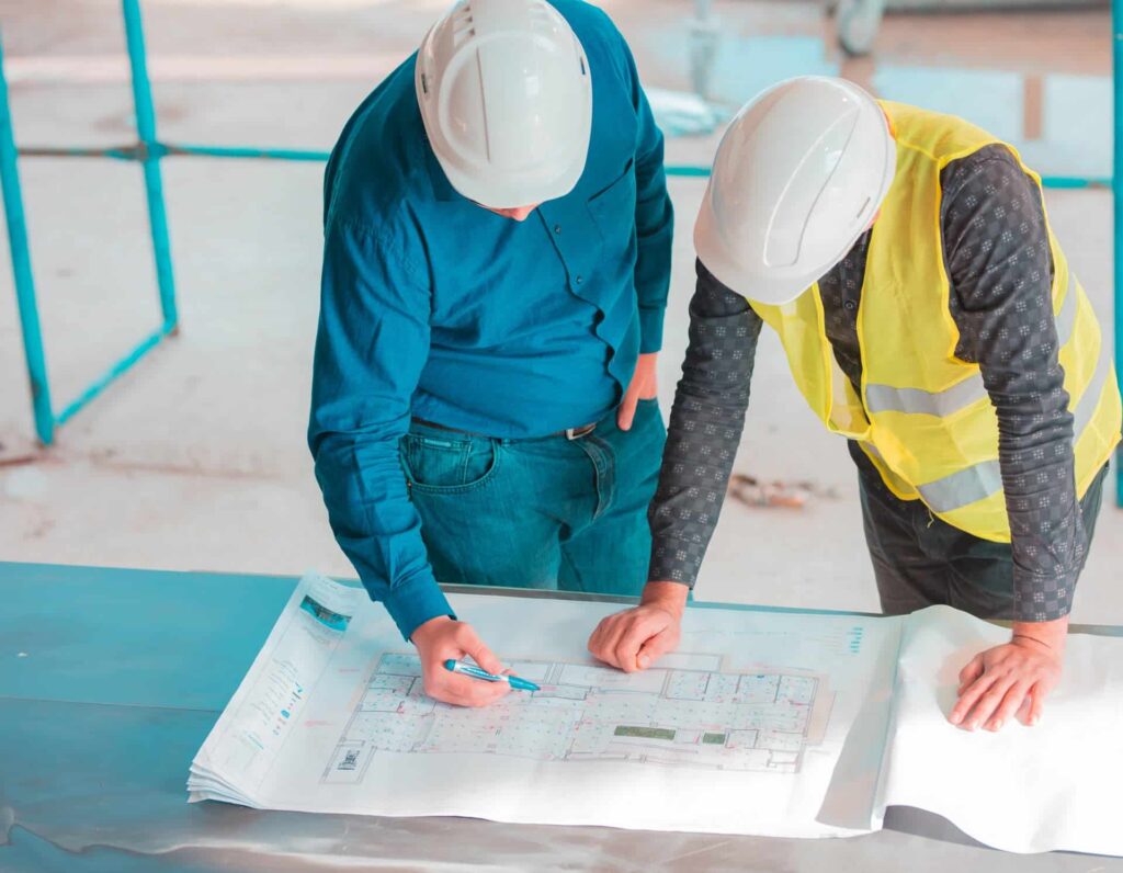 Deux personnes portant des casques de sécurité examinent des plans architecturaux sur une table sur un chantier de construction ; l'une porte un gilet de sécurité et l'autre pointe les plans à l'aide d'un stylo.