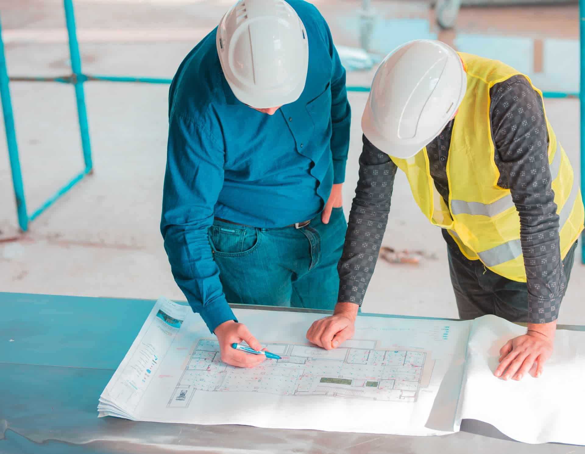Deux personnes portant des casques de sécurité examinent des plans de construction sur une table sur un chantier de construction ; l'une d'elles pointe les plans avec un stylo.
