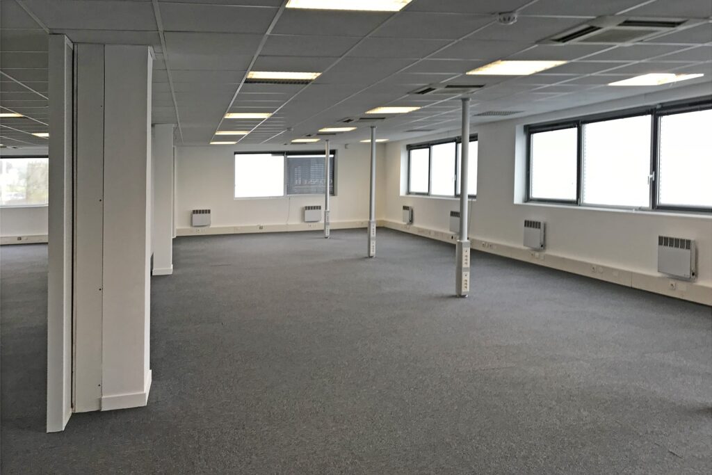 Un bureau vide avec une moquette grise, des murs blancs, des dalles de plafond blanches et de grandes fenêtres.