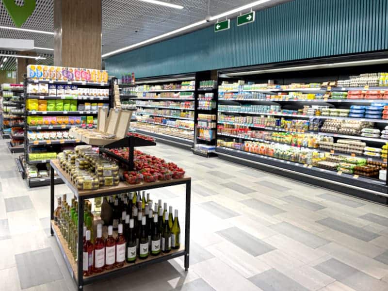 Allée d'un supermarché dont les rayons sont garnis de divers aliments emballés, de produits laitiers et de boissons ; au premier plan, une table de présentation contient des bouteilles et des bocaux de vin.