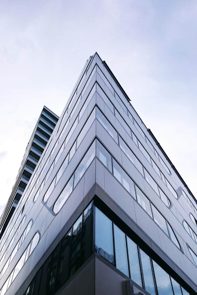 Un immeuble de bureaux moderne en verre et en métal, aux lignes anguleuses et aux fenêtres réfléchissantes, photographié depuis un angle bas et ascendant sur fond de ciel nuageux.