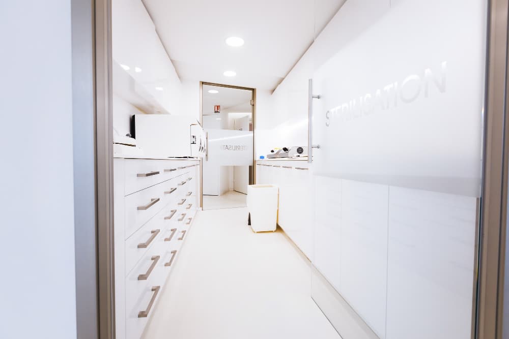 Une salle de stérilisation propre et moderne avec des armoires, des comptoirs et des tiroirs blancs, éclairés par des lampes au plafond. Une poubelle est placée près du centre.