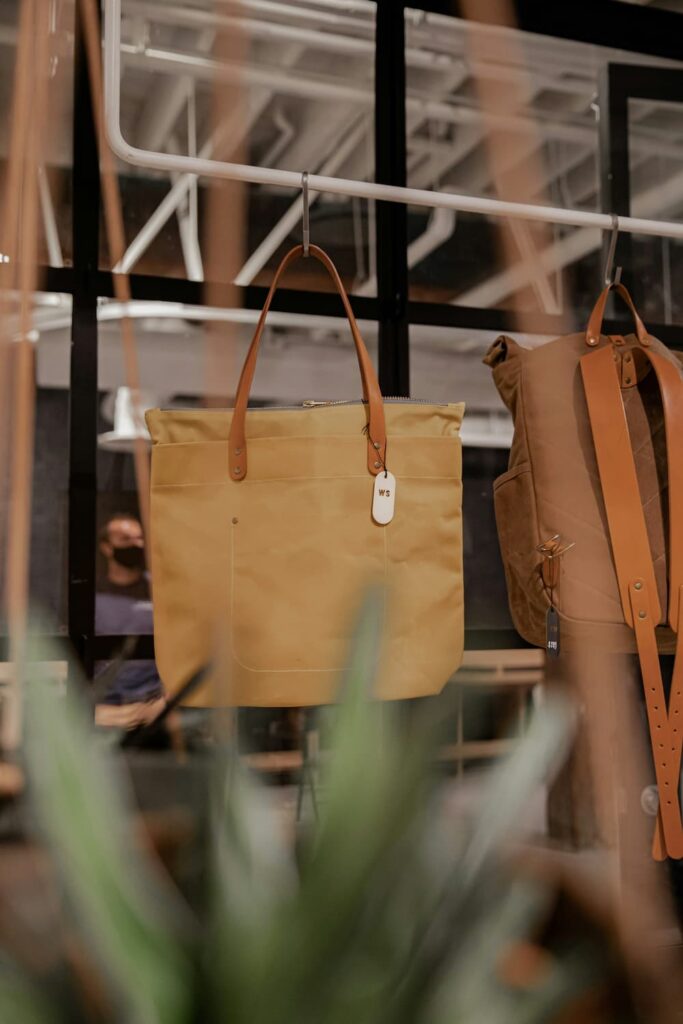 Un sac fourre-tout beige avec des poignées en cuir marron est présenté suspendu à l'intérieur, à côté d'un autre sac marron, avec une plante au premier plan.