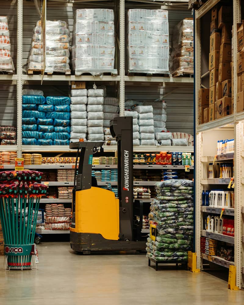 Un chariot élévateur est stationné dans une allée de la quincaillerie, entouré d'étagères remplies de sacs d'aliments pour animaux, d'outils de jardinage et de fournitures diverses.