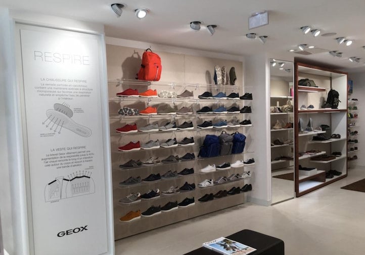 Intérieur d'un magasin de chaussures moderne avec des étagères présentant diverses chaussures de sport, des sacs à dos et des accessoires ; une affiche d'information sur la technologie des chaussures est visible sur le mur de gauche.