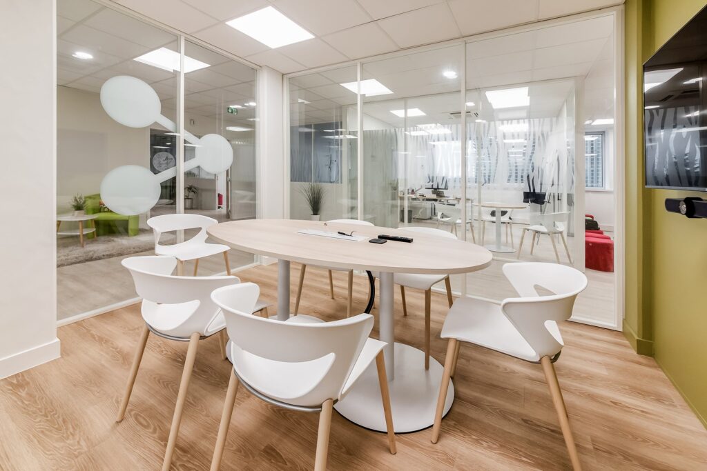 Salle de réunion moderne avec une table ronde, six chaises blanches, des murs en verre et un sol en bois. Des bureaux sont visibles à l'arrière-plan à travers les cloisons en verre.
