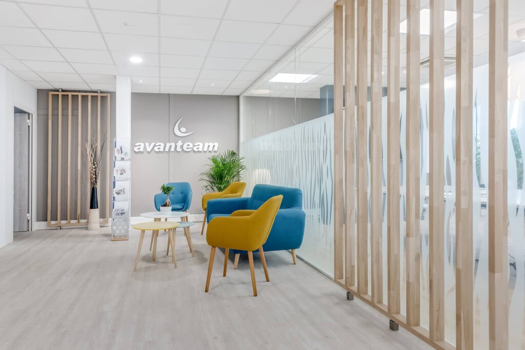 Salle d'attente du bureau avec des chaises bleues et jaunes, une cloison en lattes de bois et du verre dépoli.