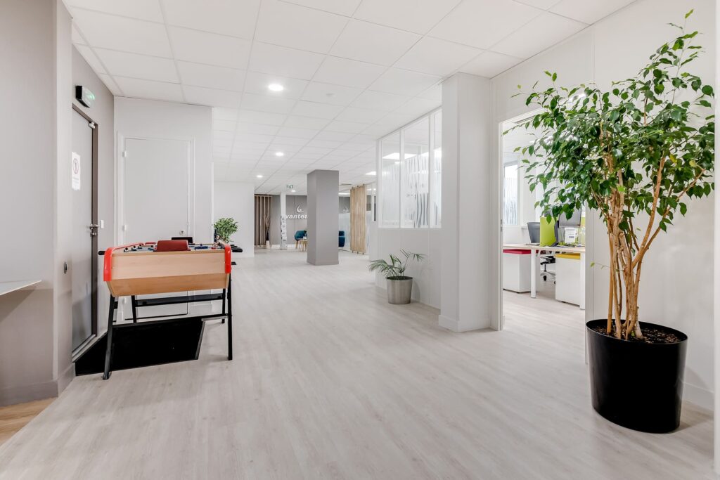 Couloir de bureau moderne avec un sol en bois clair, une plante en pot, un baby-foot et des portes ouvertes menant à des espaces de travail avec des bureaux et des chaises.