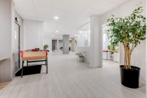 Couloir de bureau moderne avec un sol en bois clair, une plante en pot, un baby-foot et des portes ouvertes menant à des espaces de travail avec des bureaux et des chaises.