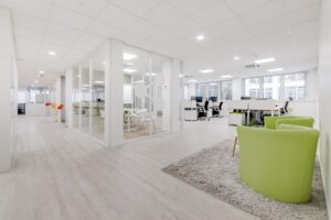 Bureau moderne à aire ouverte avec salle de réunion vitrée, bureaux blancs, chaises vertes sur un tapis gris, et lumière naturelle provenant de grandes fenêtres.