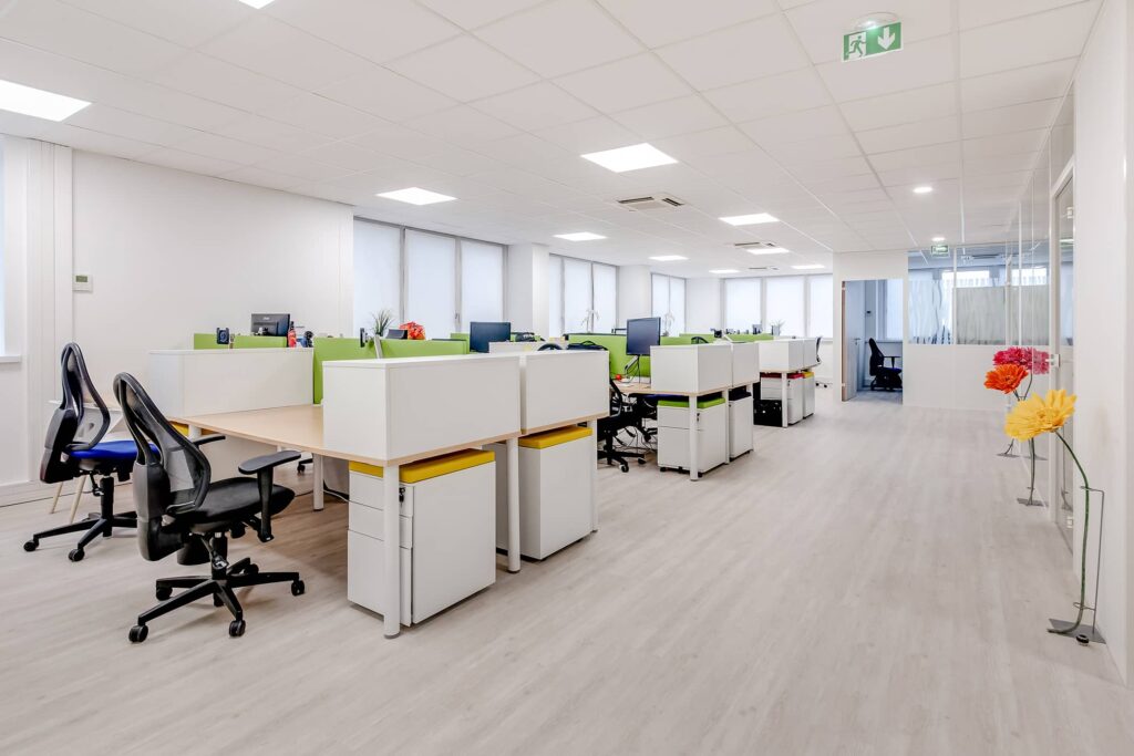 Bureau moderne à aire ouverte avec des bureaux vides, des chaises de bureau, des ordinateurs, des murs blancs, un sol en bois clair et des fleurs artificielles colorées exposées près de la cloison en verre.