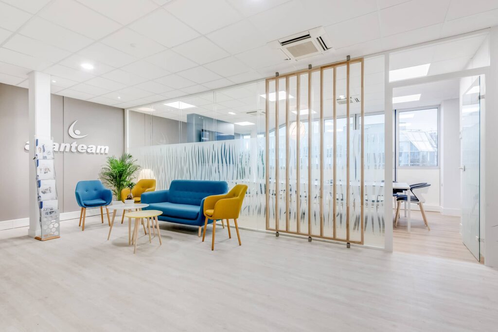 Salle d'attente d'un bureau moderne avec des chaises bleues et jaunes, de petites tables, une plante en pot et des cloisons en verre dépoli à côté d'une salle de conférence et de grandes fenêtres.