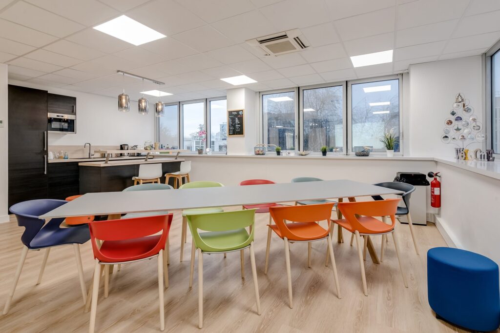 Une cuisine moderne et une salle à manger avec une longue table entourée de chaises colorées, de grandes fenêtres et une kitchenette à l'arrière-plan.