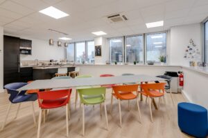 Une cuisine moderne et une salle à manger avec une longue table entourée de chaises colorées, de grandes fenêtres et une kitchenette à l'arrière-plan.