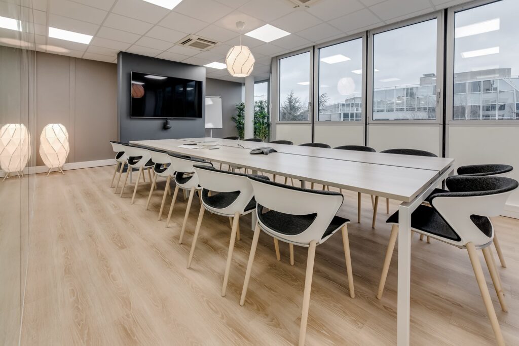 Salle de conférence moderne avec une longue table, douze chaises, un grand écran, des plafonniers, un sol en bois et des fenêtres donnant sur des immeubles de bureaux.