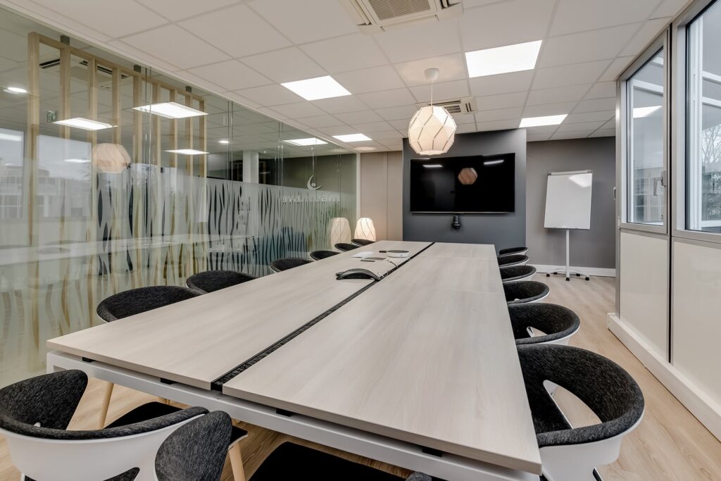 Salle de conférence moderne équipée d'une longue table de réunion, de chaises noires, d'un grand écran mural, d'un tableau à feuilles mobiles et de parois en verre. La salle est dotée d'un sol en bois clair et d'un éclairage contemporain.