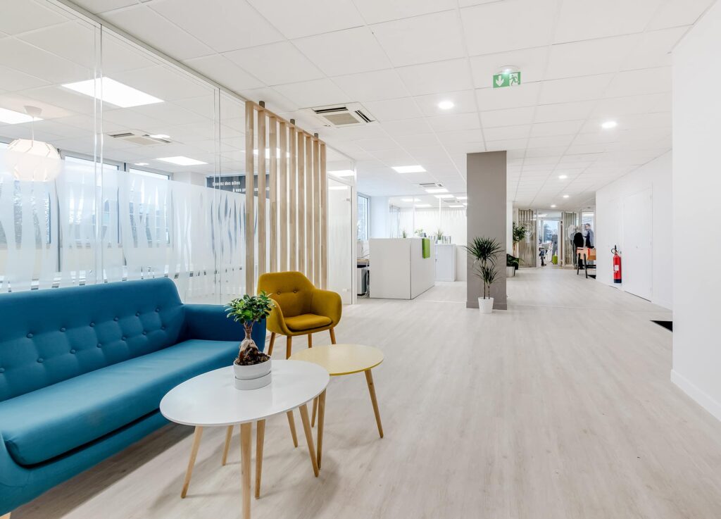 Couloir de bureau moderne avec un sol en bois clair, des murs en verre, un canapé bleu, un fauteuil jaune, des tables blanches, des plantes en pot et des bureaux en arrière-plan.