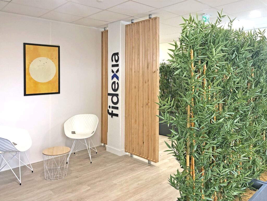 Salle d'attente d'un bureau moderne avec deux chaises blanches, une petite table ronde, des plantes en pot, une cloison en bois, des œuvres d'art murales et le mot "fidexia" affiché verticalement.