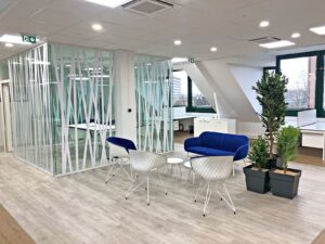 Espace de bureau moderne avec salle de réunion vitrée, canapé bleu, chaises blanches, petites tables, plantes en pot et grandes fenêtres offrant une lumière naturelle.