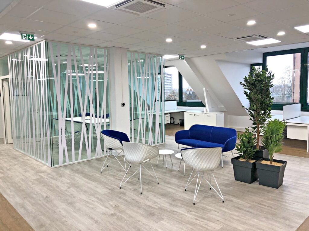 Espace de bureau moderne avec une salle de réunion en verre, un canapé bleu, des chaises blanches, de petites tables, des plantes en pot, un sol en bois et de grandes fenêtres.