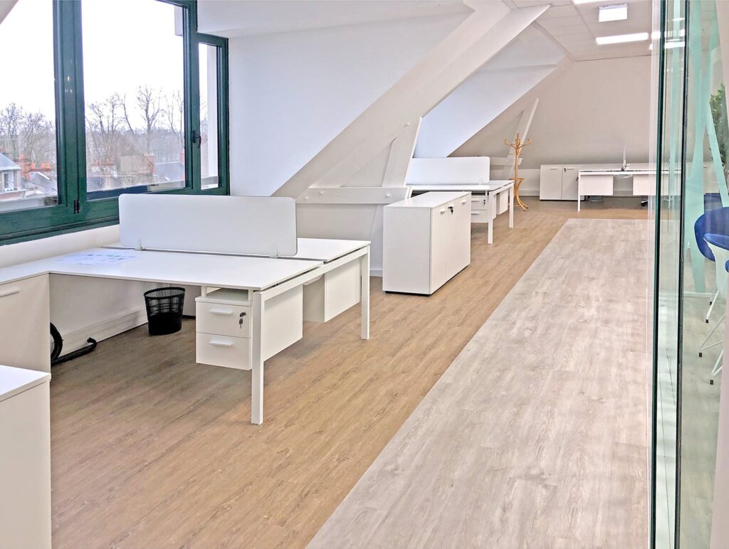 Un bureau moderne avec plusieurs bureaux, chaises et armoires blancs disposés le long d'une pièce lumineuse avec de grandes fenêtres et un sol en bois.