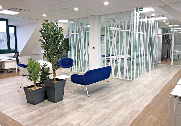Espace de bureau moderne avec salle de réunion vitrée, sièges bleus et blancs, plantes d'intérieur, sol en bois et lumière naturelle grâce à de grandes fenêtres.