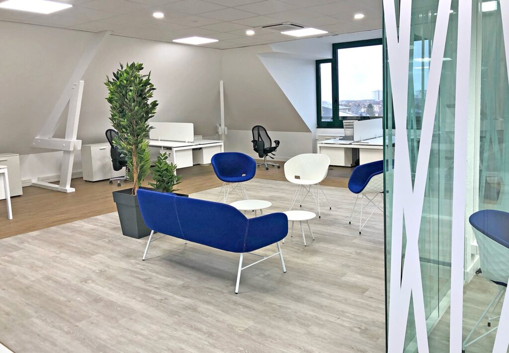 Bureau moderne avec des bureaux blancs, des chaises noires, un petit coin salon avec des sièges bleus et blancs, une plante en pot et de grandes fenêtres qui apportent de la lumière naturelle.