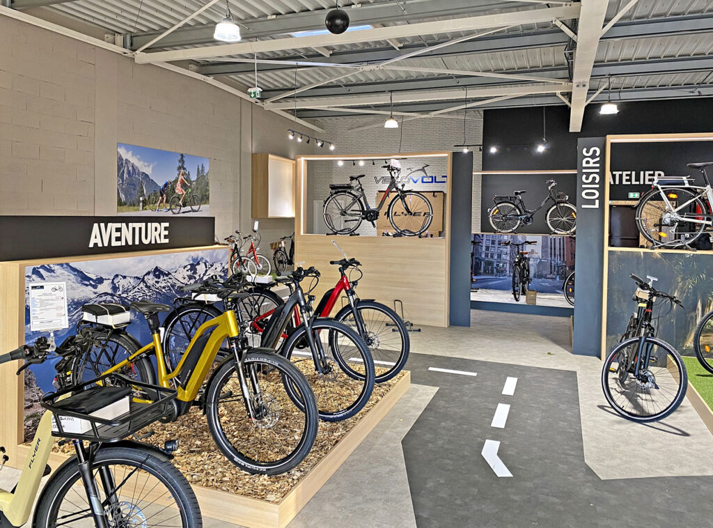 Une salle d'exposition présente divers vélos sur des supports, avec des panneaux indiquant "Aventure", "Loisirs" et "Atelier". De grandes photos de scènes extérieures ornent les murs.