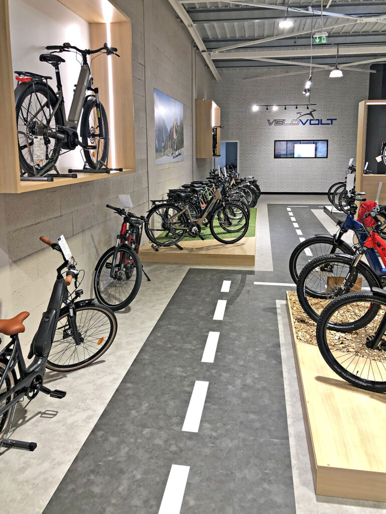 Des rangées de bicyclettes sont exposées à l'intérieur d'un magasin de vélos moderne, avec un faux motif de route sur le sol et des bicyclettes montées au mur et sur des supports au sol.