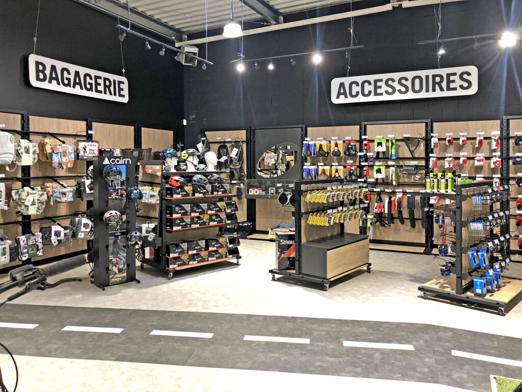 Intérieur d'un magasin de détail présentant divers sacs, bagages et accessoires sur des étagères et des rayonnages, avec des sections étiquetées "Bagagerie" et "Accessoires" sur les murs.