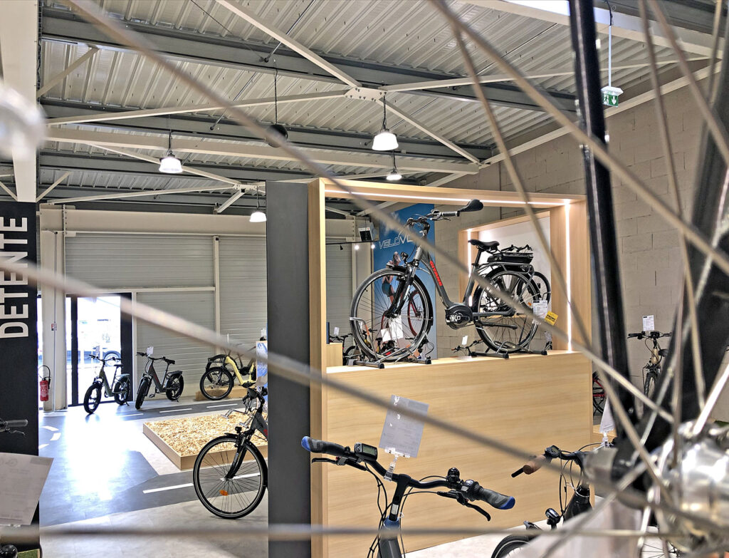 Bicyclettes exposées à l'intérieur d'une salle d'exposition moderne dotée de grandes fenêtres et d'un éclairage zénithal, vues à travers les rayons d'une roue de bicyclette au premier plan.