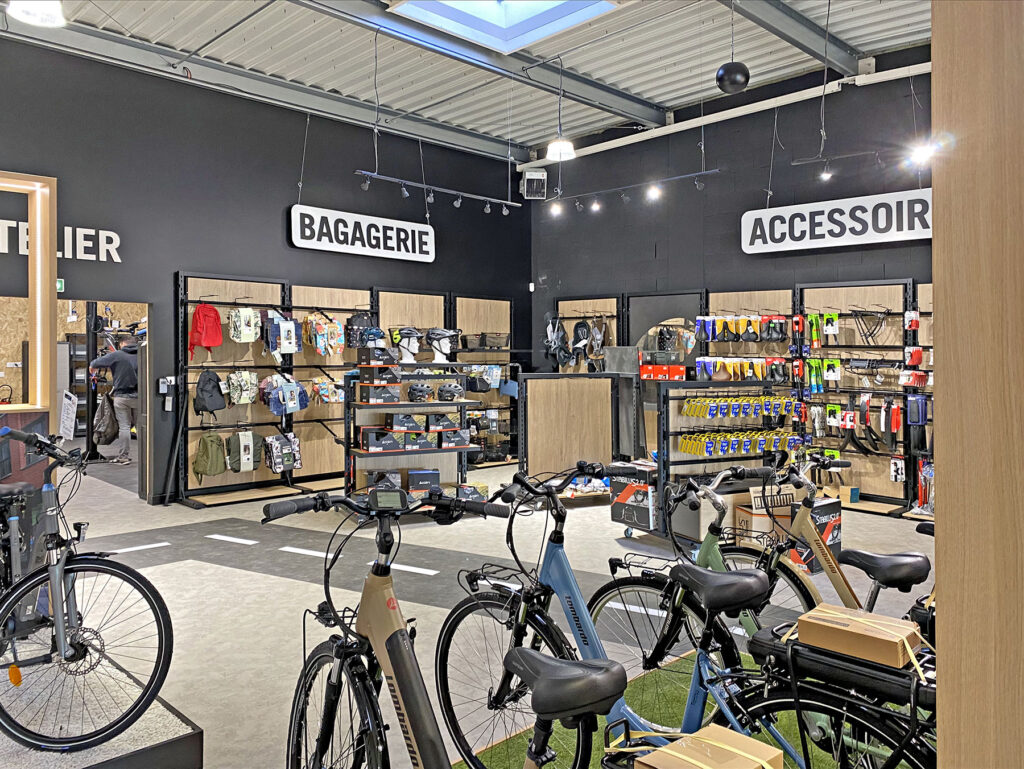 Intérieur d'un magasin de bicyclettes avec des vélos exposés au premier plan et des étagères de sacs et d'accessoires contre des murs noirs portant les inscriptions "BAGAGERIE" et "ACCESSOIRE".
