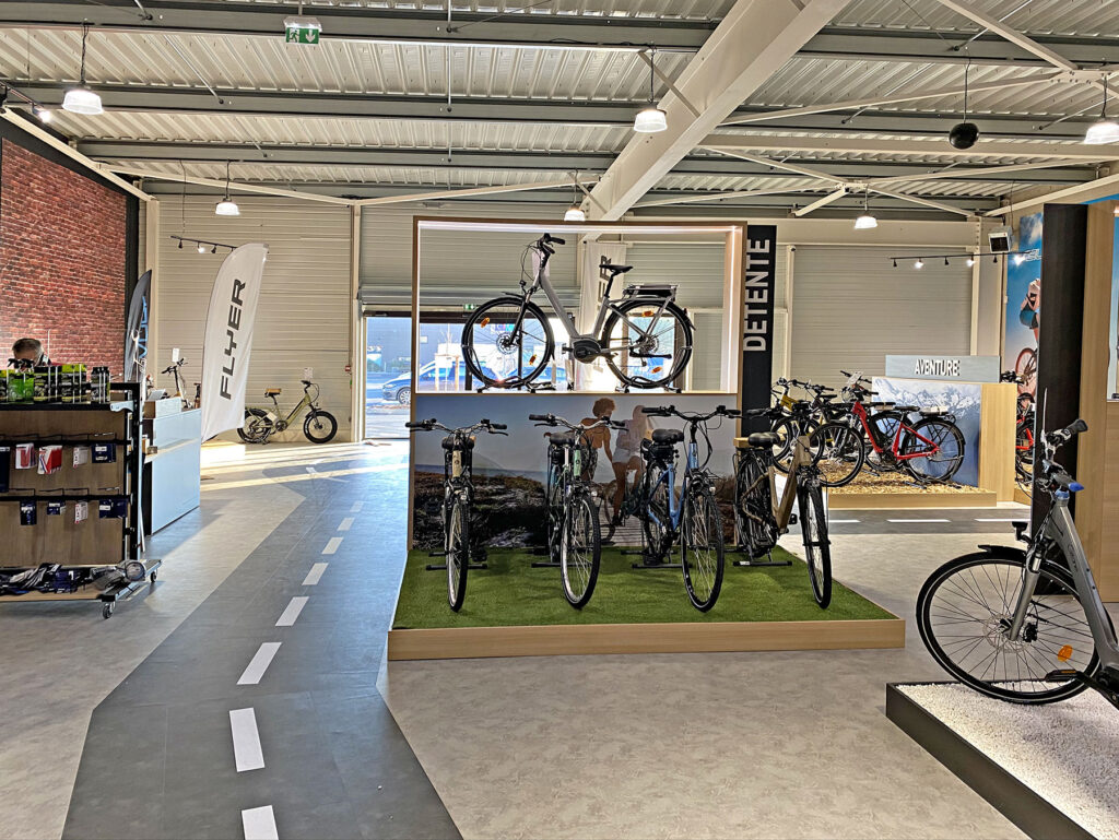 Les vélos sont exposés sur des supports dans une salle d'exposition bien éclairée, avec un sol à motifs routiers et divers accessoires de cyclisme sur des étagères.