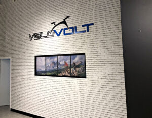 Un mur de briques blanches affiche le logo "VELOVOLT" au-dessus d'une fenêtre présentant une photo panoramique de vélo de montagne.