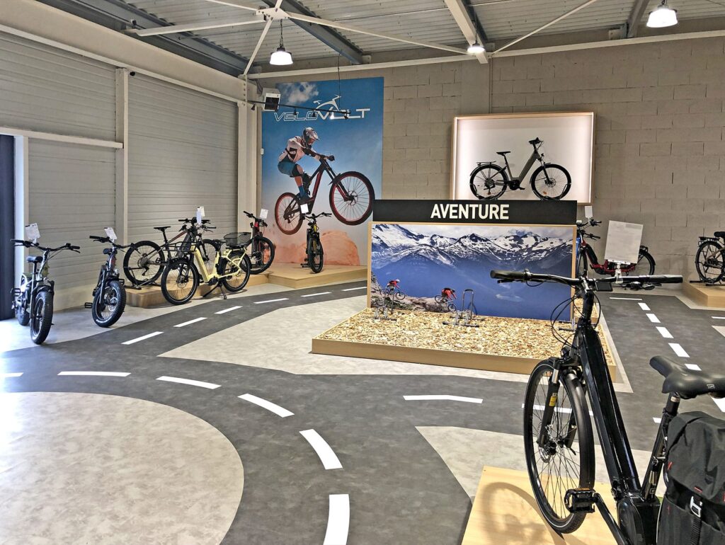 Salle d'exposition intérieure de bicyclettes avec divers vélos exposés, des marquages routiers sur le sol et des affiches sur le thème de l'aventure sur les murs.