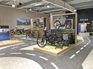 Une salle d'exposition de vélos moderne présente divers vélos sur des plates-formes et des supports au sol, avec en arrière-plan des simulations de marquage routier et une zone étiquetée "Atelier".