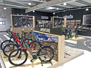 Intérieur d'un magasin de bicyclettes avec des vélos de montagne en exposition, des supports en bois étiquetés "Aventure" et divers accessoires de cyclisme sur des étagères à l'arrière-plan.