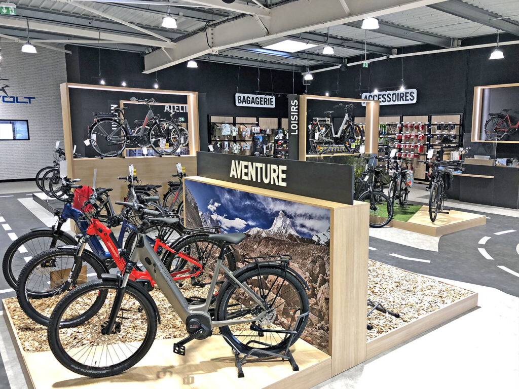Intérieur d'un magasin de vélos avec plusieurs vélos exposés, y compris des modèles électriques, et des sections étiquetées pour les accessoires, les bagages et les articles de loisir.