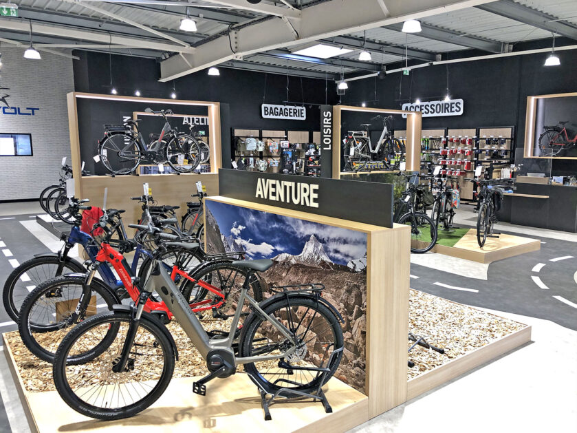 L'intérieur d'un magasin de vélos moderne présente divers vélos, accessoires et équipements sur des racks et des étagères, avec des panneaux en français indiquant les différentes sections.