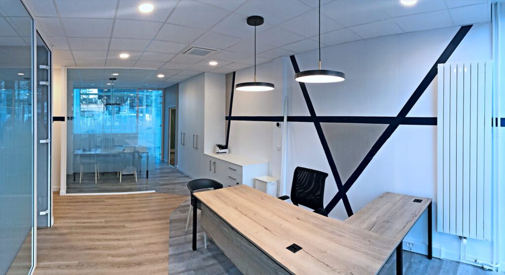Bureau moderne avec un bureau en bois, une chaise noire, des murs géométriques, des lampes suspendues et une salle de réunion vitrée en arrière-plan.