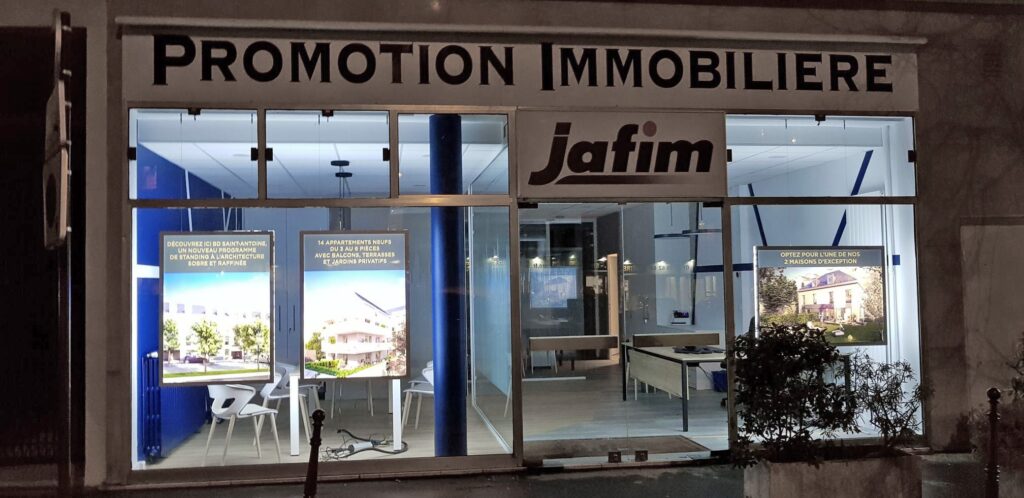 Façade d'une agence immobilière nommée "Promotion Immobilière Jafim" de nuit, avec des annonces immobilières illuminées visibles à travers les vitres.