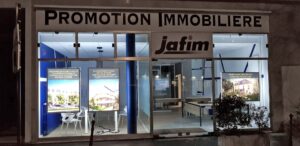 La façade d'une agence immobilière la nuit, avec des enseignes lumineuses affichant des annonces immobilières et le nom "jafim" au-dessus de l'entrée.