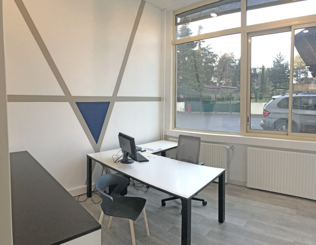 Bureau moderne avec deux bureaux, deux écrans et des chaises près d'une grande fenêtre ; décor minimaliste avec des murs géométriques et un sol en bois.