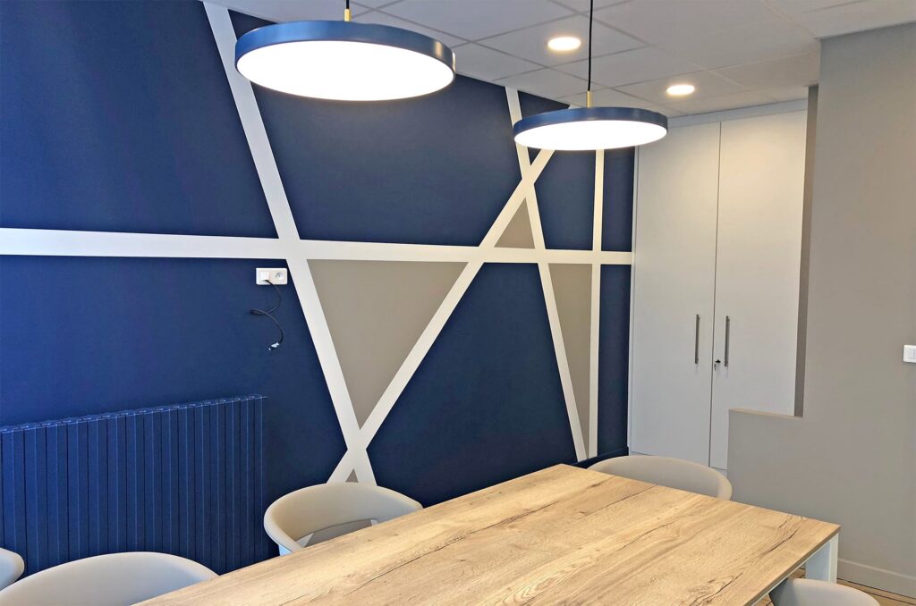 Une salle de réunion moderne avec une table en bois, des chaises beiges, un mur d'accent géométrique bleu, deux plafonniers ronds et des armoires blanches encastrées.