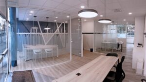 Espace de bureau moderne avec salles de réunion vitrées, parquet en bois clair, murs blancs et mobilier minimaliste, avec des plafonniers ronds et de grandes fenêtres.