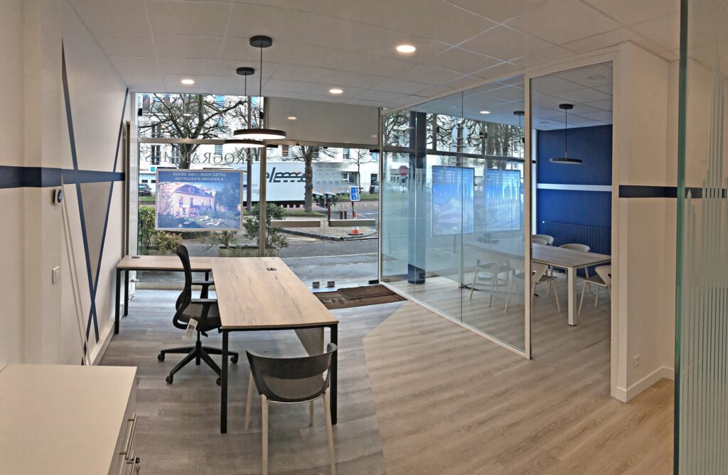 Bureau moderne avec de grandes fenêtres, deux bureaux, des chaises noires et une salle de réunion vitrée avec une table et des chaises blanches. Vue de la rue urbaine visible à l'extérieur.