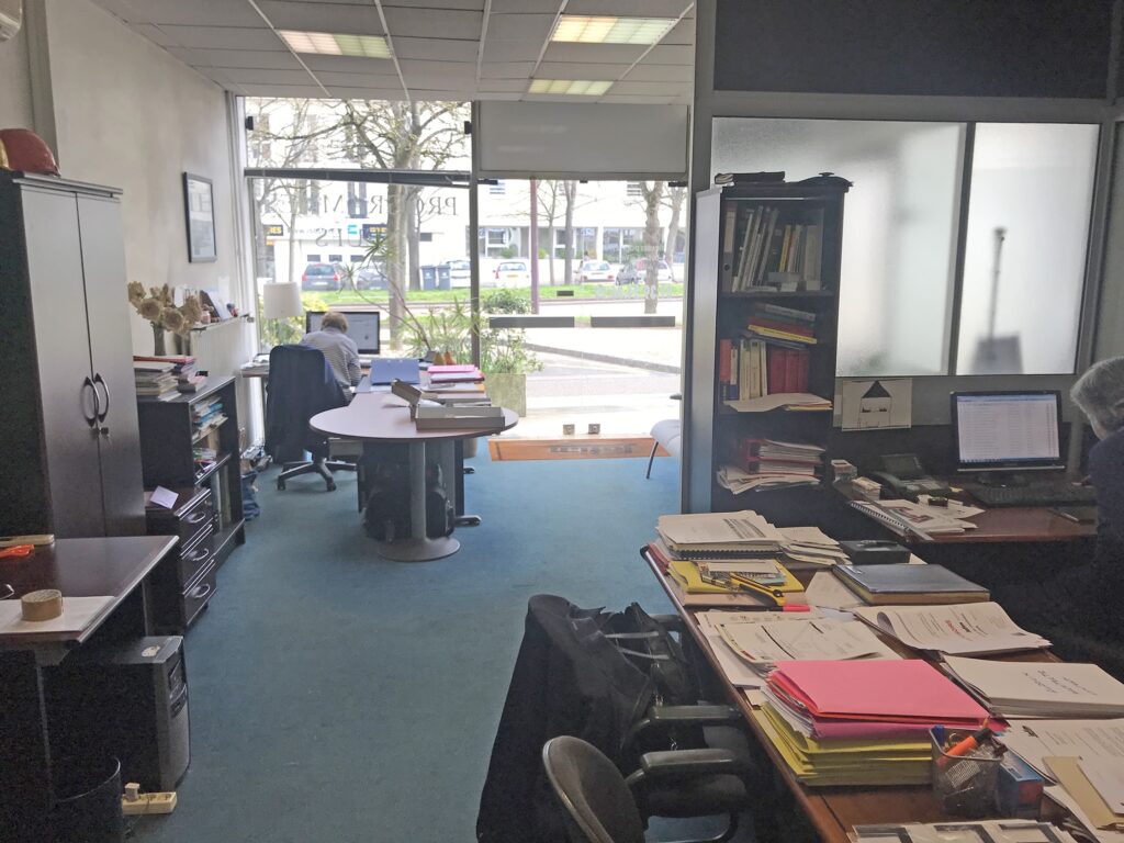 Bureau avec deux bureaux, des piles de documents, des étagères et une grande fenêtre donnant sur la rue ; une personne est assise et travaille à un bureau près de la fenêtre.