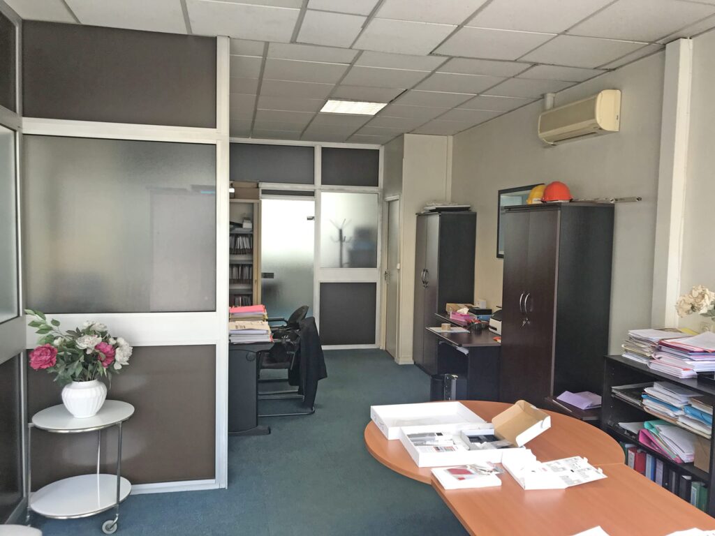 Bureau avec des bureaux, des chaises, des armoires, des piles de papiers et une plante en pot ; l'éclairage fluorescent et les cloisons en verre dépoli sont visibles.